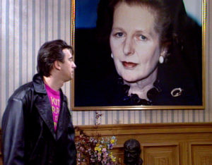 Kit, in a leather jacket, finds a giant portrait of Margaret Thatcher