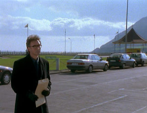 John (wearing glasses) holds an envelope as he approaches a hotel