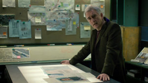 Steve standing at centre table over a case file with a concerned expression