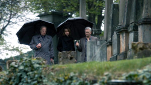 Steve shares an umbrella with MacDougall. Gerry walks besides them. They're in a graveyard.